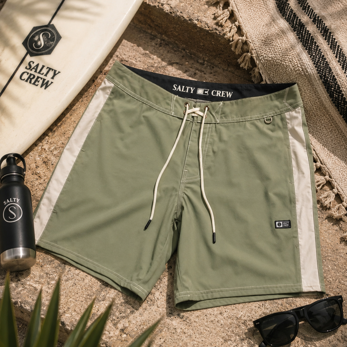 Bañador Salty Crew Channel Boardshort Oil Green para hombre sobre fondo lifestyle surf con tabla de surf y accesorios premium de playa