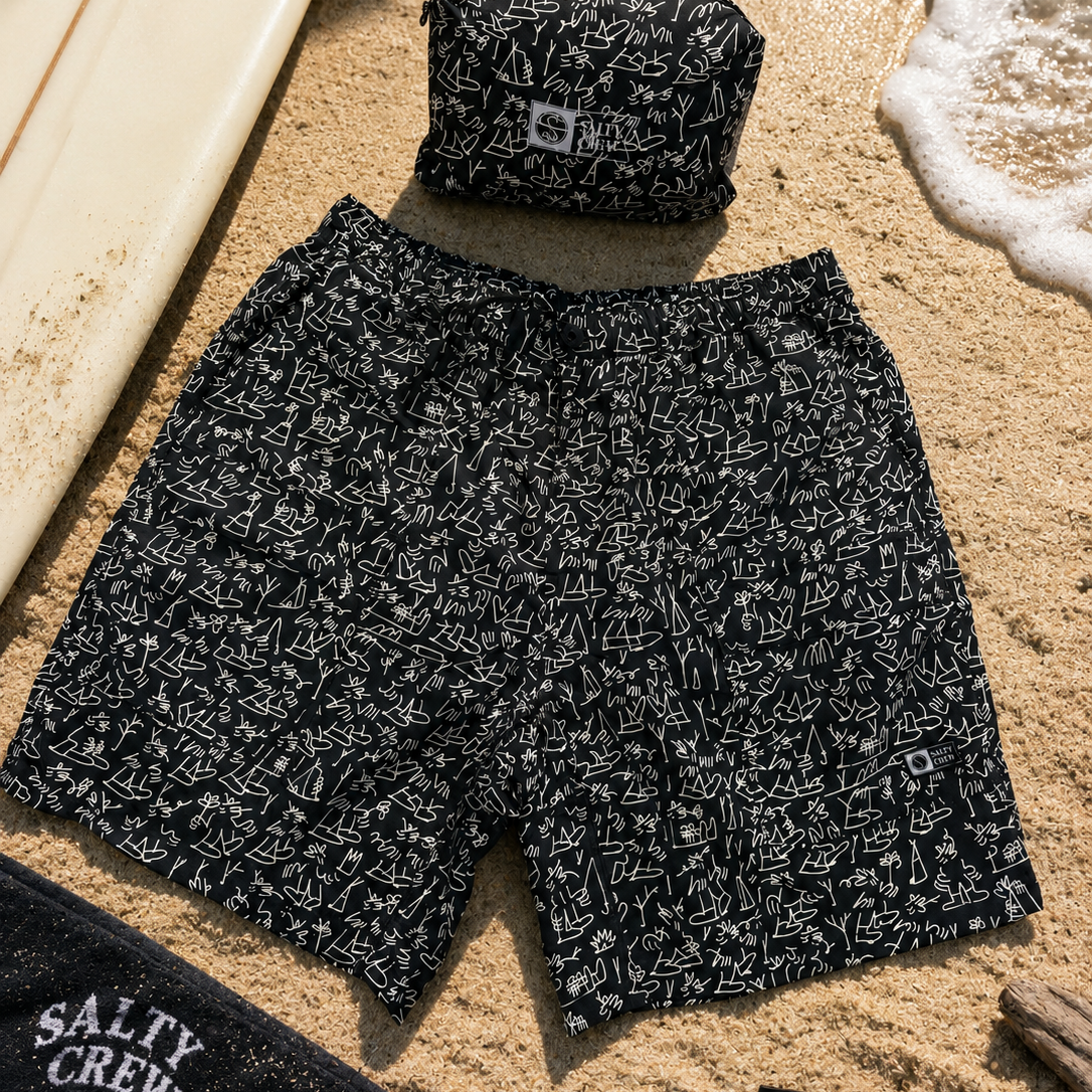 Bañador Salty Crew Raid Packable Short LB Black negro con estampado surfero sobre arena de playa junto a tabla de surf y accesorios lifestyle