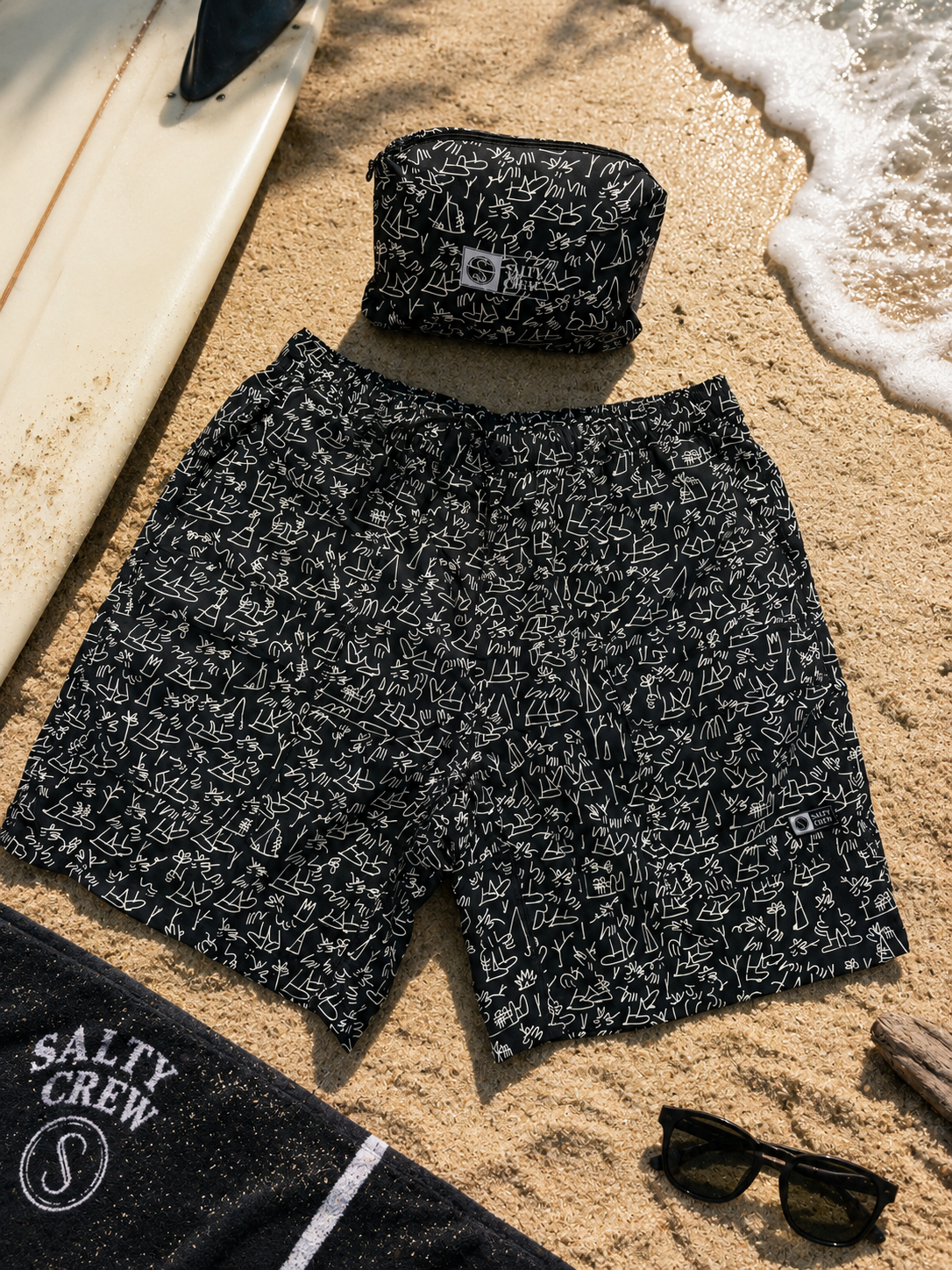 Bañador Salty Crew Raid Packable Short LB Black negro con estampado surfero sobre arena de playa junto a tabla de surf y accesorios lifestyle