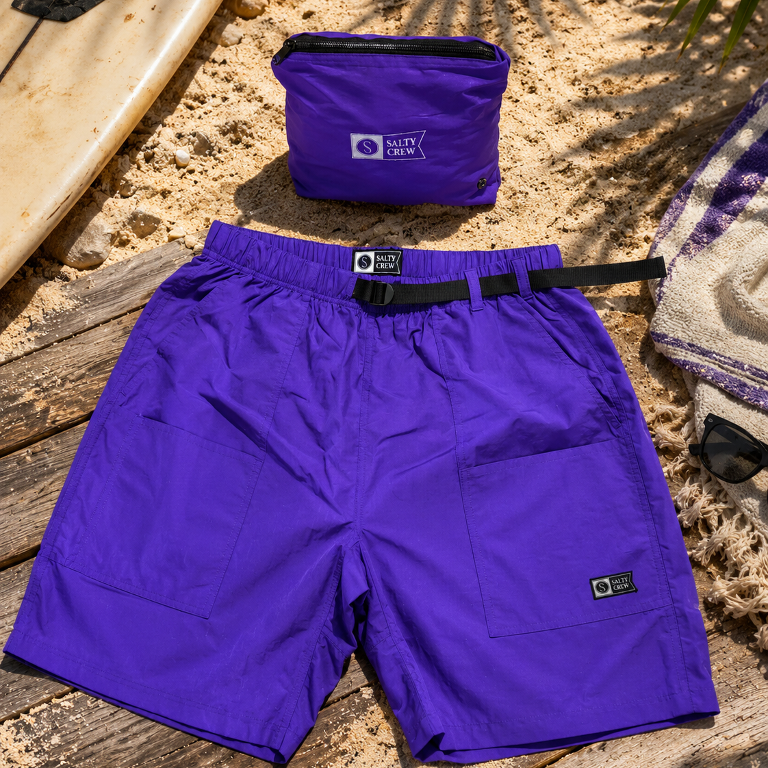 Bañador Salty Crew Raid Packable Short Purple Heart morado sobre escenario lifestyle de playa con tabla de surf y accesorios surferos para hombre