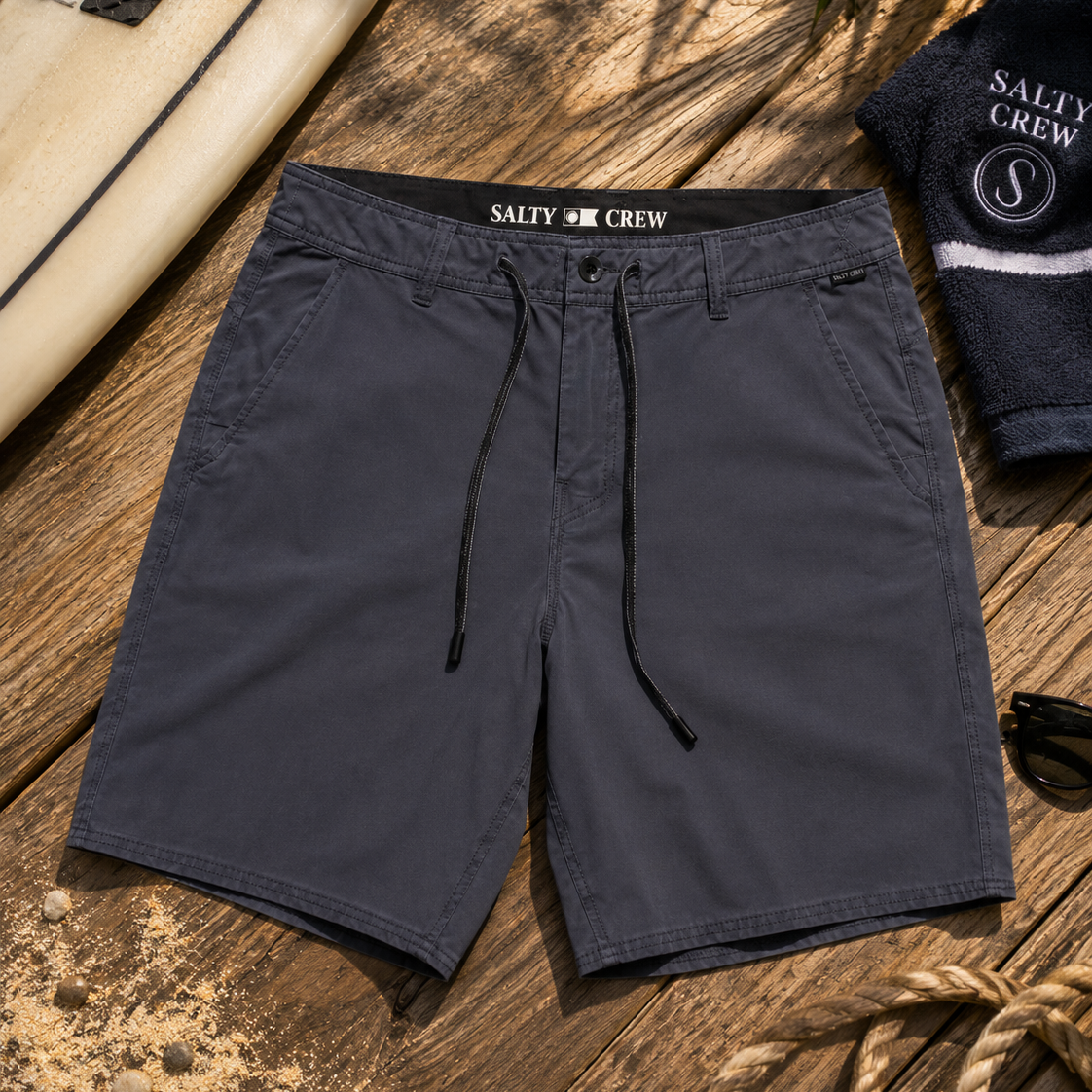 Bermudas híbridas Salty Crew Transom Overdye Phantom para hombre sobre fondo lifestyle de playa con tabla de surf y accesorios surferos premium
