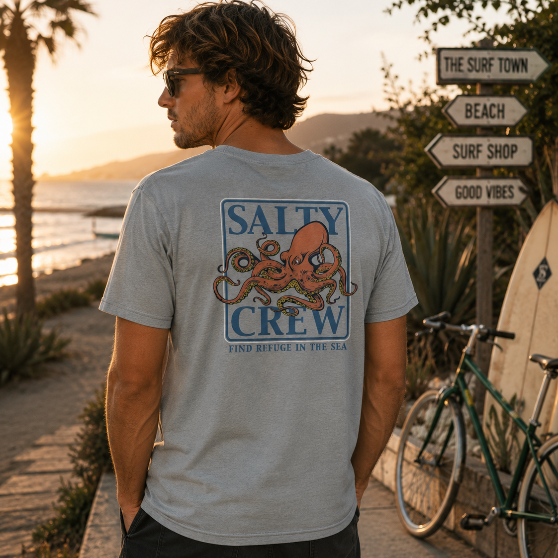 Camiseta hombre Salty Crew pulpo Ink Slinger Tee color quarry en paseo marítimo surf lifestyle premium con cartel The Surf Town al atardecer Costa del Sol