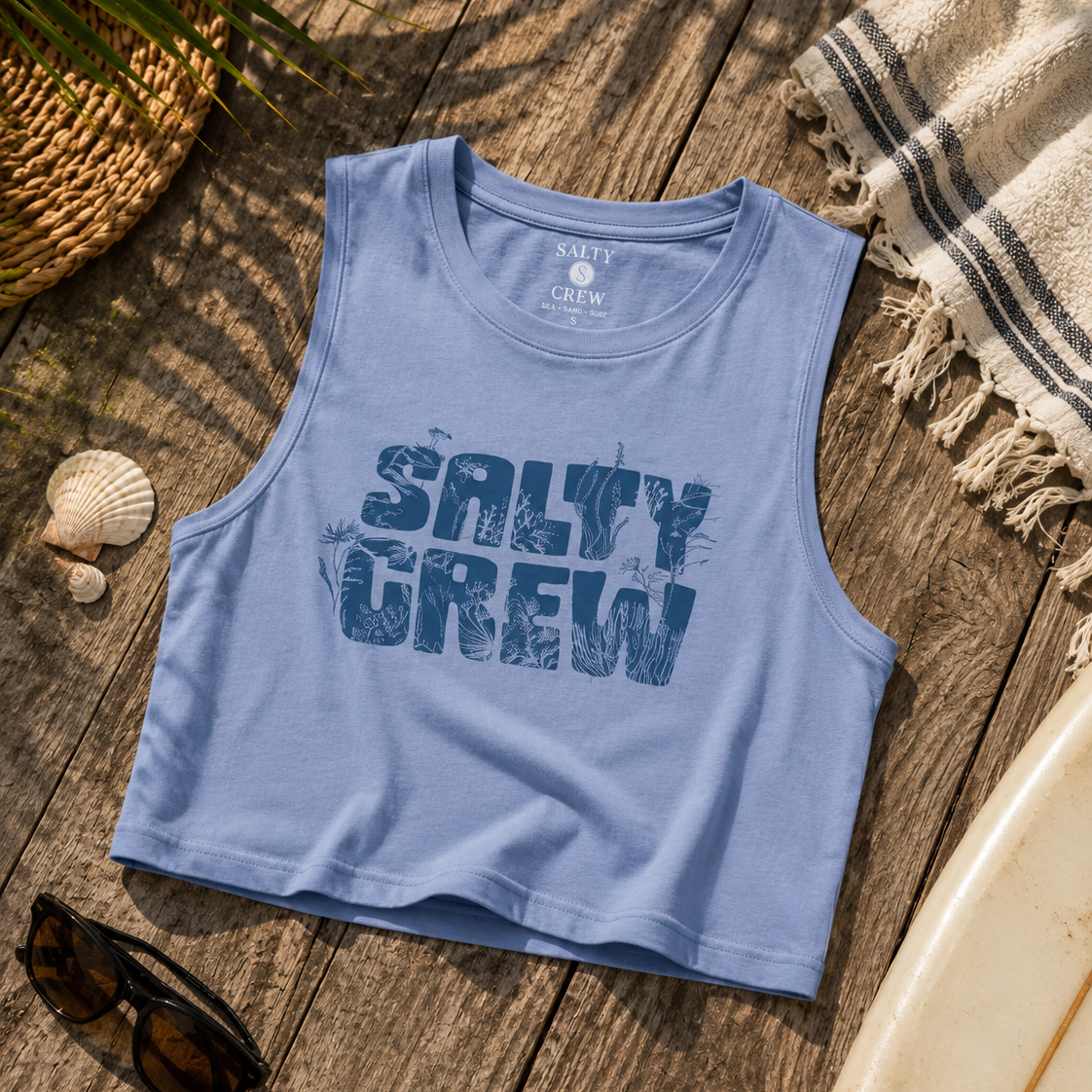 Camiseta cropped Salty Crew Outside Tank Violet Blue para mujer sobre fondo lifestyle de playa con tabla de surf y accesorios surferos premium