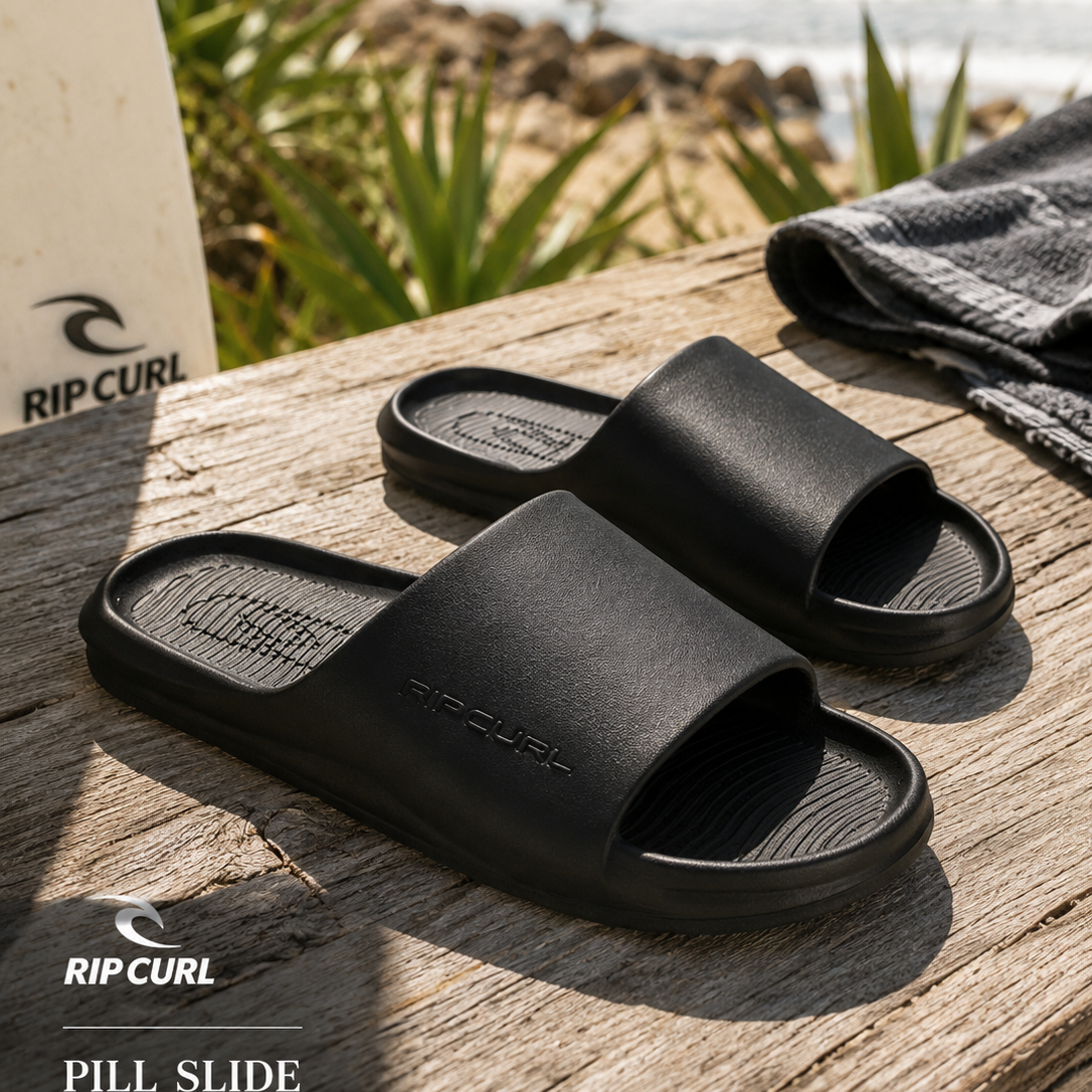 Chanclas de pala Rip Curl Pill Slide Black en escena lifestyle surf sobre madera frente al mar con luz natural de verano