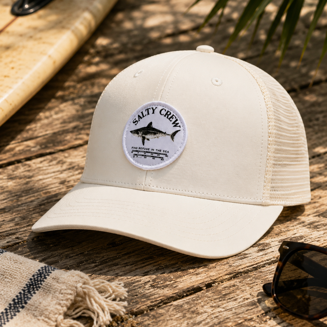 Gorra Salty Crew Bruce Trucker Off White sobre fondo lifestyle de playa con tabla de surf y accesorios surferos premium