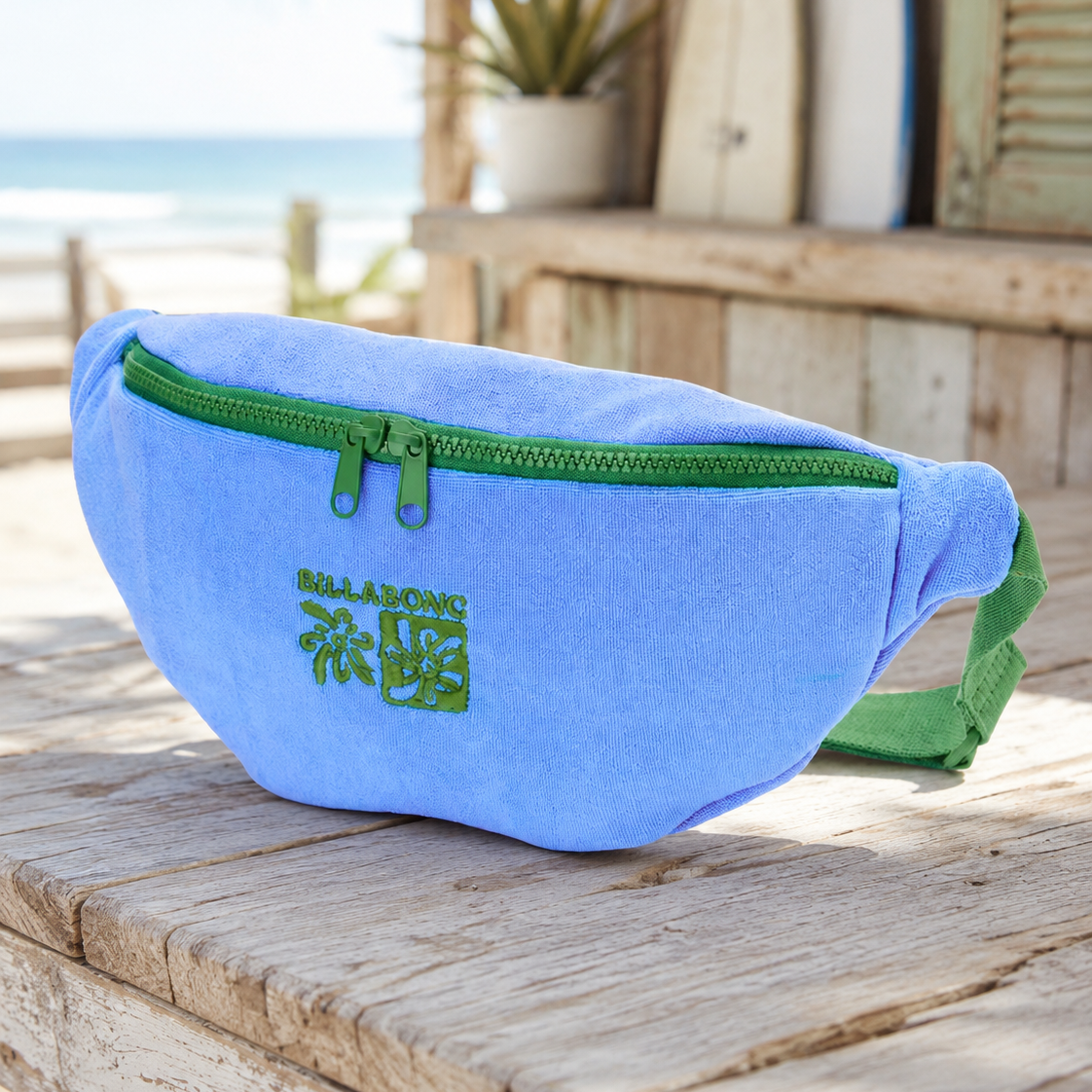 Riñonera Billabong Long Weekend True Blue azul con cremallera verde y logo bordado, bolso de cintura estilo surf lifestyle sobre mesa de madera con ambiente playa.