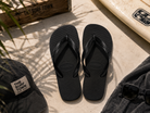 Chanclas Havaianas Top negras sobre suelo mediterráneo junto a tabla de surf y accesorios surferos premium con detalle The Surf Town Surf Shop