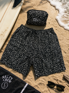 Bañador Salty Crew Raid Packable Short LB Black negro con estampado surfero sobre arena de playa junto a tabla de surf y accesorios lifestyle