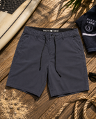Bermudas híbridas Salty Crew Transom Overdye Phantom para hombre sobre fondo lifestyle de playa con tabla de surf y accesorios surferos premium