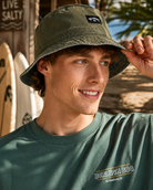 Sombrero bucket reversible Billabong verde estilo surf lifestyle en escena editorial premium junto al mar