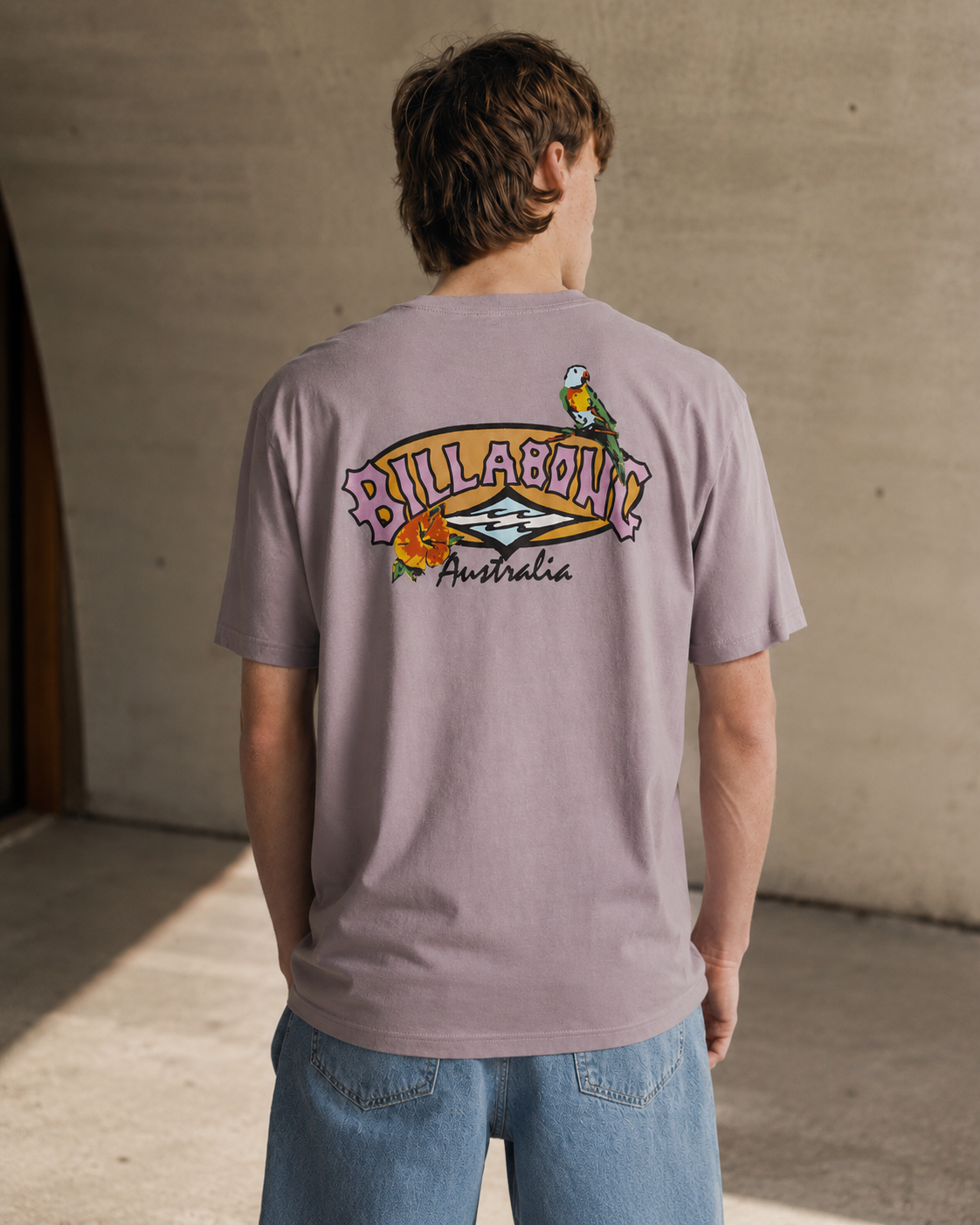 Camiseta Billabong Origin Purple Ash para hombre con estampado tropical trasero en escena editorial surf premium de estilo minimalista y luz cálida natural.