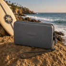 Bolsito Havaianas Mini Bag gris oscuro sobre rocas frente al mar junto a tabla de surf en ambiente coastal lifestyle premium