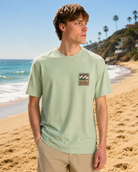 Camiseta Billabong verde hombre estilo surf premium en playa soleada frente al mar
