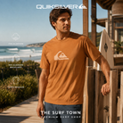 Hombre con camiseta Quiksilver Comp Logo Tobacco Brown frente al mar, imagen editorial propia de THE SURF TOWN