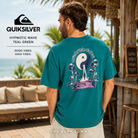 Camiseta Quiksilver Hypnotic Wave Teal Green para hombre en beach club tropical de Bali, fotografía editorial propia de THE SURF TOWN, camiseta verde turquesa con gráfico trasero psicodélico inspirado en el surf y el verano.