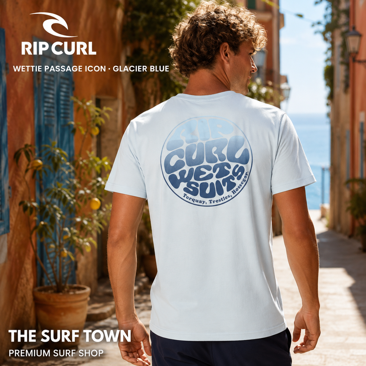 Camiseta Rip Curl Wettie Passage Icon Tee Glacier Blue para hombre mostrando el gráfico trasero azul en una calle mediterránea, imagen editorial propia de THE SURF TOWN.