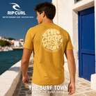 Camiseta Rip Curl Wettie Passage Icon Tee Mustard Gold para hombre mostrando el gráfico trasero crema junto a un puerto mediterráneo, imagen editorial propia de THE SURF TOWN.