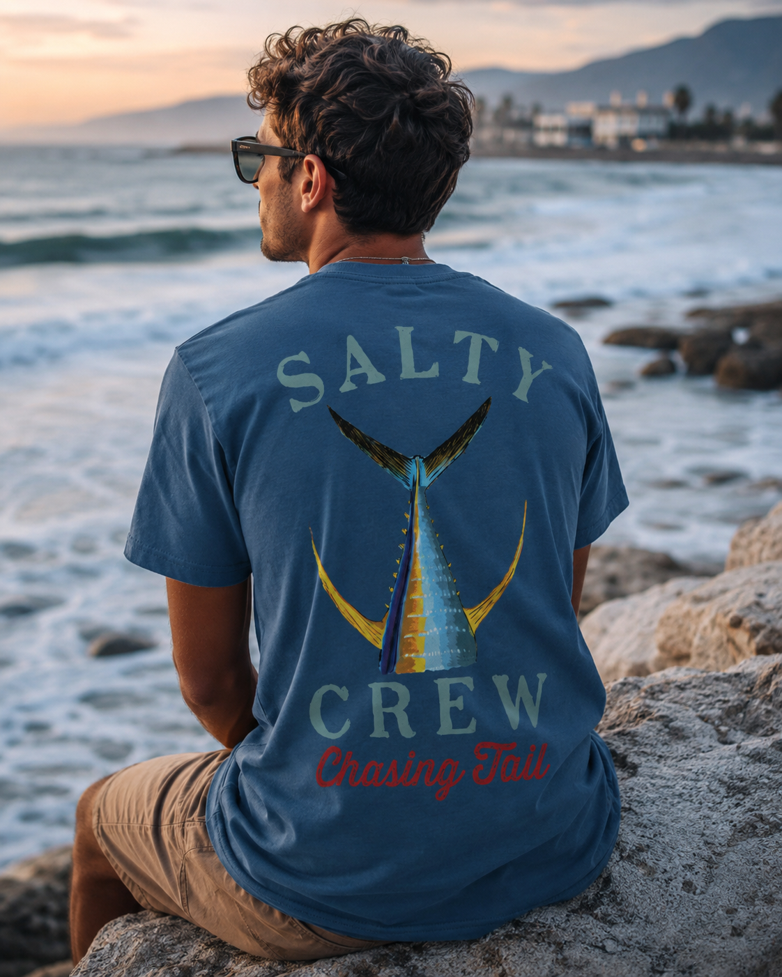 Camiseta Salty Crew Chasing Tail Cast Blue para hombre en escena lifestyle surf sobre rocas junto al mar al atardecer Costa del Sol