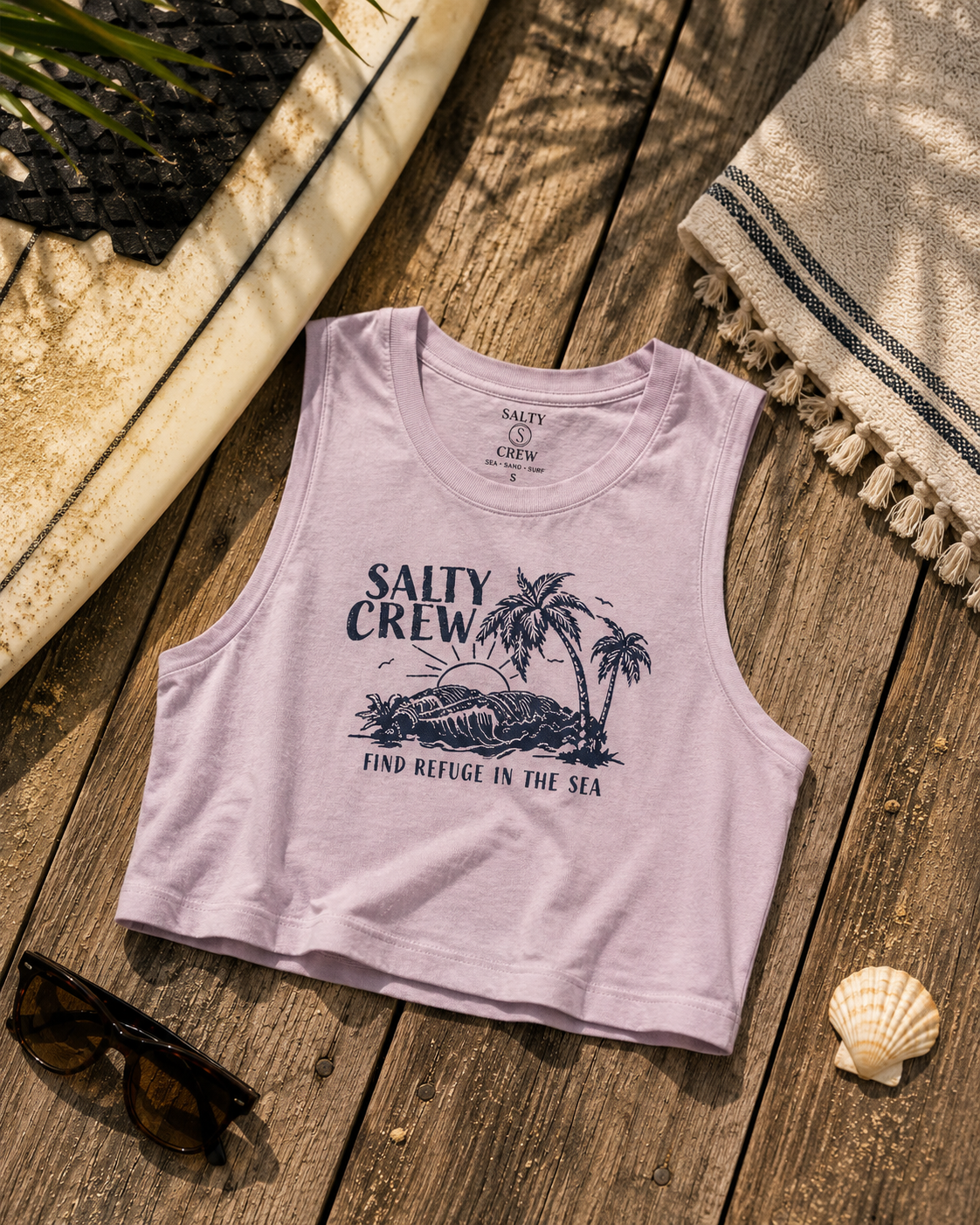 Camiseta cropped Salty Crew Outside Tank Pale Orchid para mujer sobre fondo lifestyle de playa con tabla de surf y accesorios surferos premium