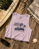 Camiseta cropped Salty Crew Outside Tank Pale Orchid para mujer sobre fondo lifestyle de playa con tabla de surf y accesorios surferos premium