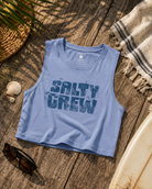 Camiseta cropped Salty Crew Outside Tank Violet Blue para mujer sobre fondo lifestyle de playa con tabla de surf y accesorios surferos premium