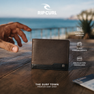 Cartera Rip Curl Ridge PU All Day Brown sobre una mesa frente al océano, imagen editorial propia de THE SURF TOWN