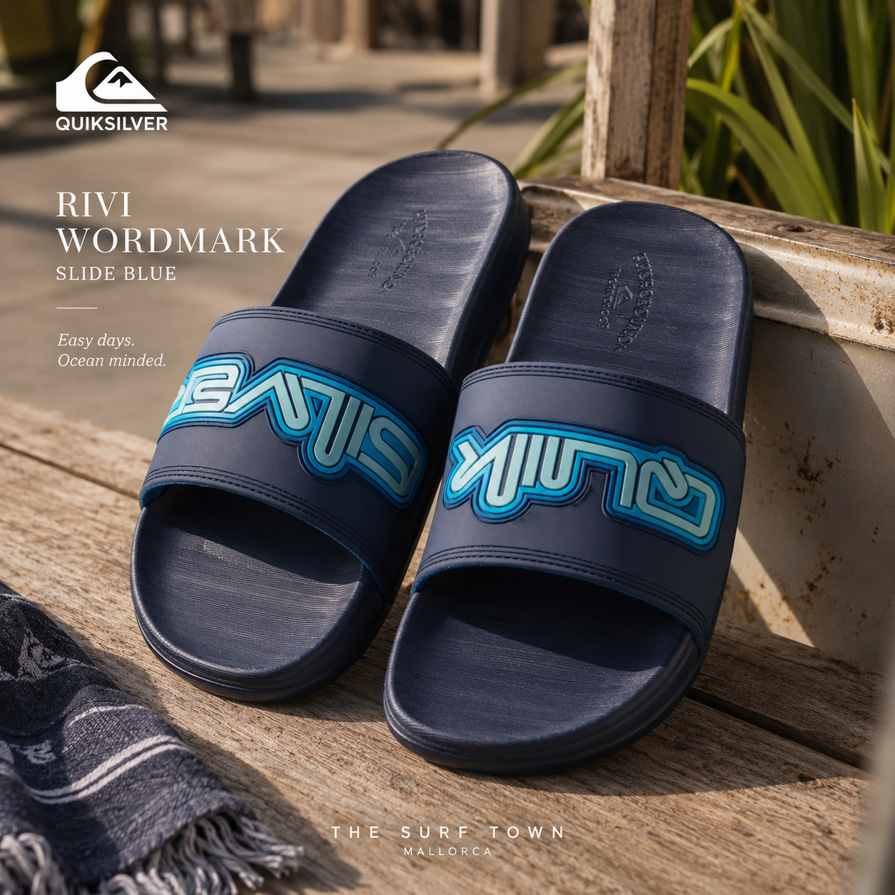 Chanclas de pala Quiksilver Rivi Wordmark Slide Blue en fotografía editorial propia de THE SURF TOWN sobre banco de madera surf con estilo coastal lifestyle y luz natural premium