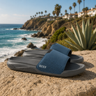 Chanclas pala Reef Tailslide Grey Navy para hombre sobre piedra junto al mar con estilo surf lifestyle premium y ambiente Costa del Sol