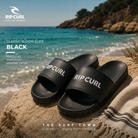 Chanclas de pala Rip Curl Classic Bloom Slide Black en fotografía editorial propia de THE SURF TOWN sobre una playa mediterránea de arena blanca
