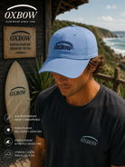 Gorra Oxbow Elini Lost Blue azul con logo frontal en escenario coastal premium y estética editorial surf cinematográfica