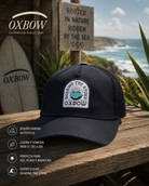 Gorra Oxbow Stoked Deep Marine azul marino con parche en escenario coastal premium y estética editorial surf cinematográfica
