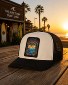 Gorra trucker Rip Curl Padang Padang blanca y negra lifestyle surf premium sobre mesa frente al mar al atardecer