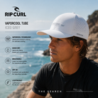 Gorra Rip Curl Vaporcool Tube Iced Grey en fotografía editorial propia de THE SURF TOWN con surfero frente al mar, tecnología VaporCool, tejido transpirable de secado rápido, protección solar UPF y estilo surf técnico para verano.