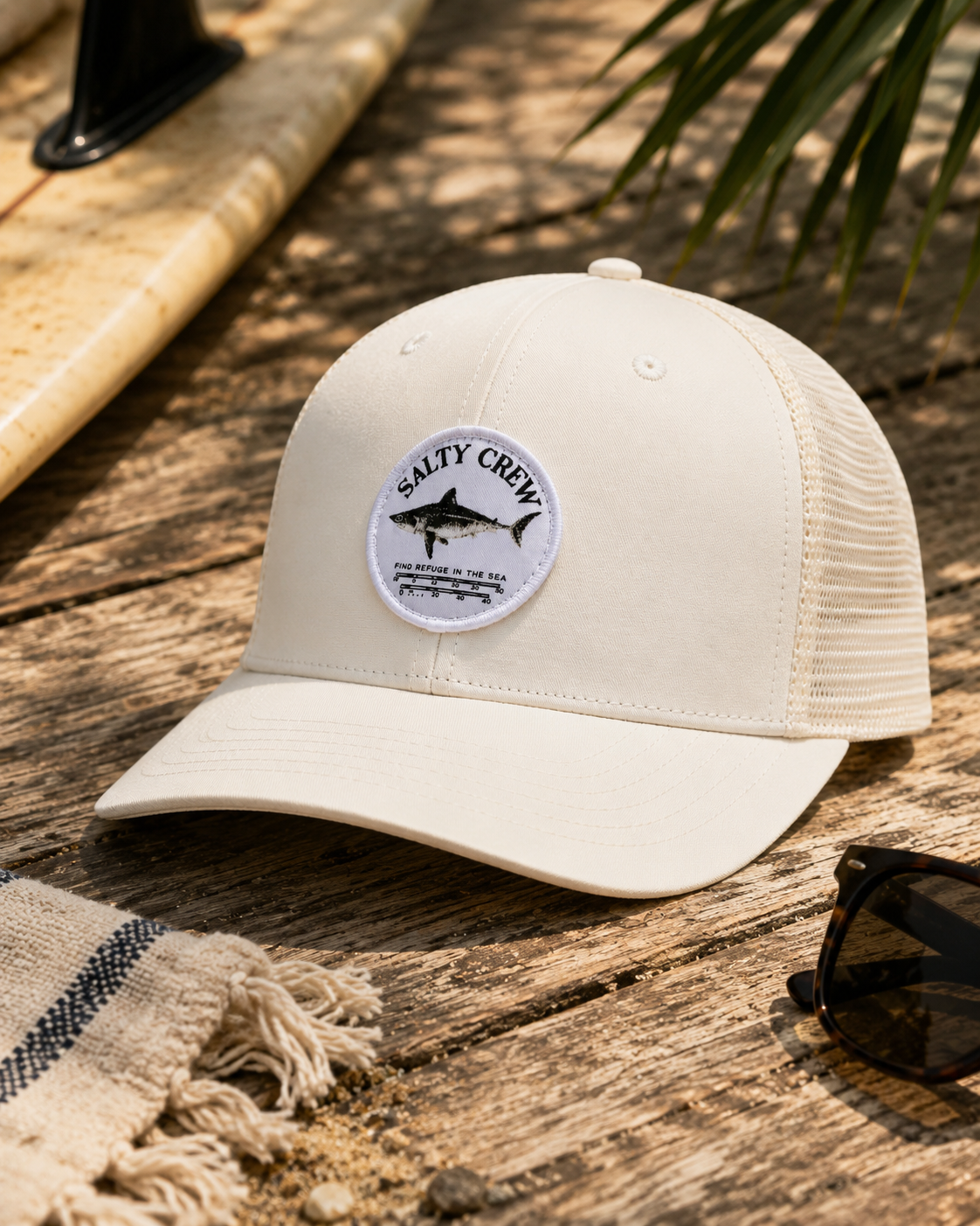 Gorra Salty Crew Bruce Trucker Off White sobre fondo lifestyle de playa con tabla de surf y accesorios surferos premium