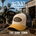 Gorra Von Dutch Used 05 en una fotografía editorial exclusiva creada por THE SURF TOWN con estética café racer, moto custom y luz natural diurna, destacando el parche bordado Von Dutch, el diseño trucker premium y la combinación de beige, azul marino y amarillo mostaza.