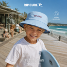 Niño con gorro Rip Curl Combo Mid Brim azul en un paseo junto a la playa, imagen editorial propia de THE SURF TOWN