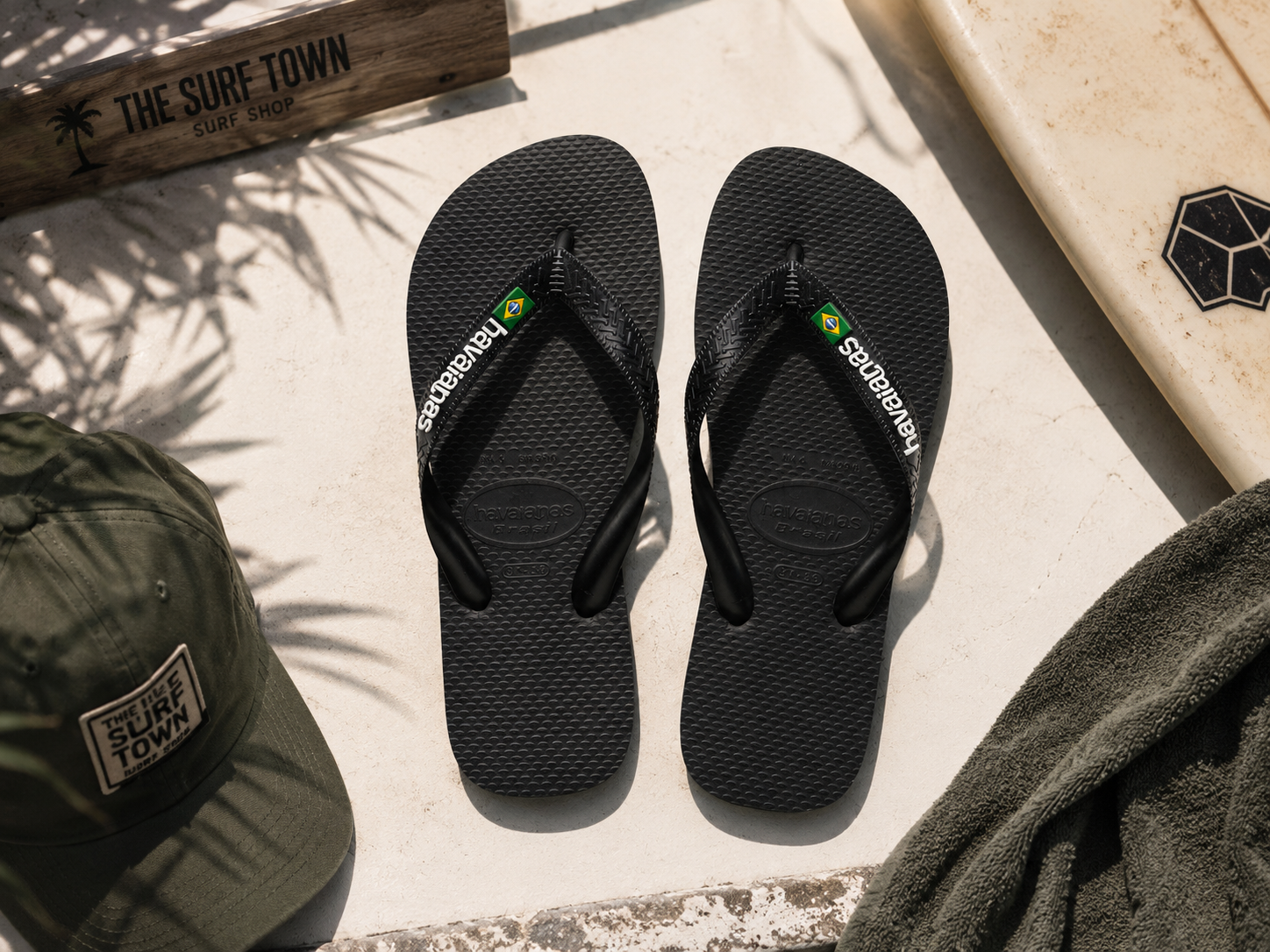 Chanclas Havaianas Brasil Logo negras sobre suelo mediterráneo junto a tabla de surf y accesorios surferos en ambiente lifestyle masculino premium con detalle The Surf Town