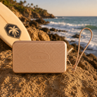 Bolsito Havaianas Mini Bag champagne sobre rocas frente al mar junto a tabla de surf en ambiente coastal lifestyle premium