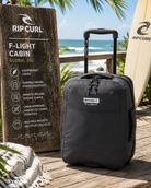 Maleta Rip Curl F-Light Cabin 30L Midnight Black en escenario surf travel premium con estética coastal adventure luminosa