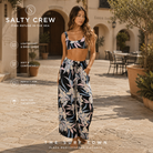 Pantalones Salty Crew Dreamland Beach Pant Black en plaza mediterránea elegante, fotografía editorial propia de THE SURF TOWN con modelo lifestyle femenino, pantalón fluido estampado tropical y estilo resort premium para verano