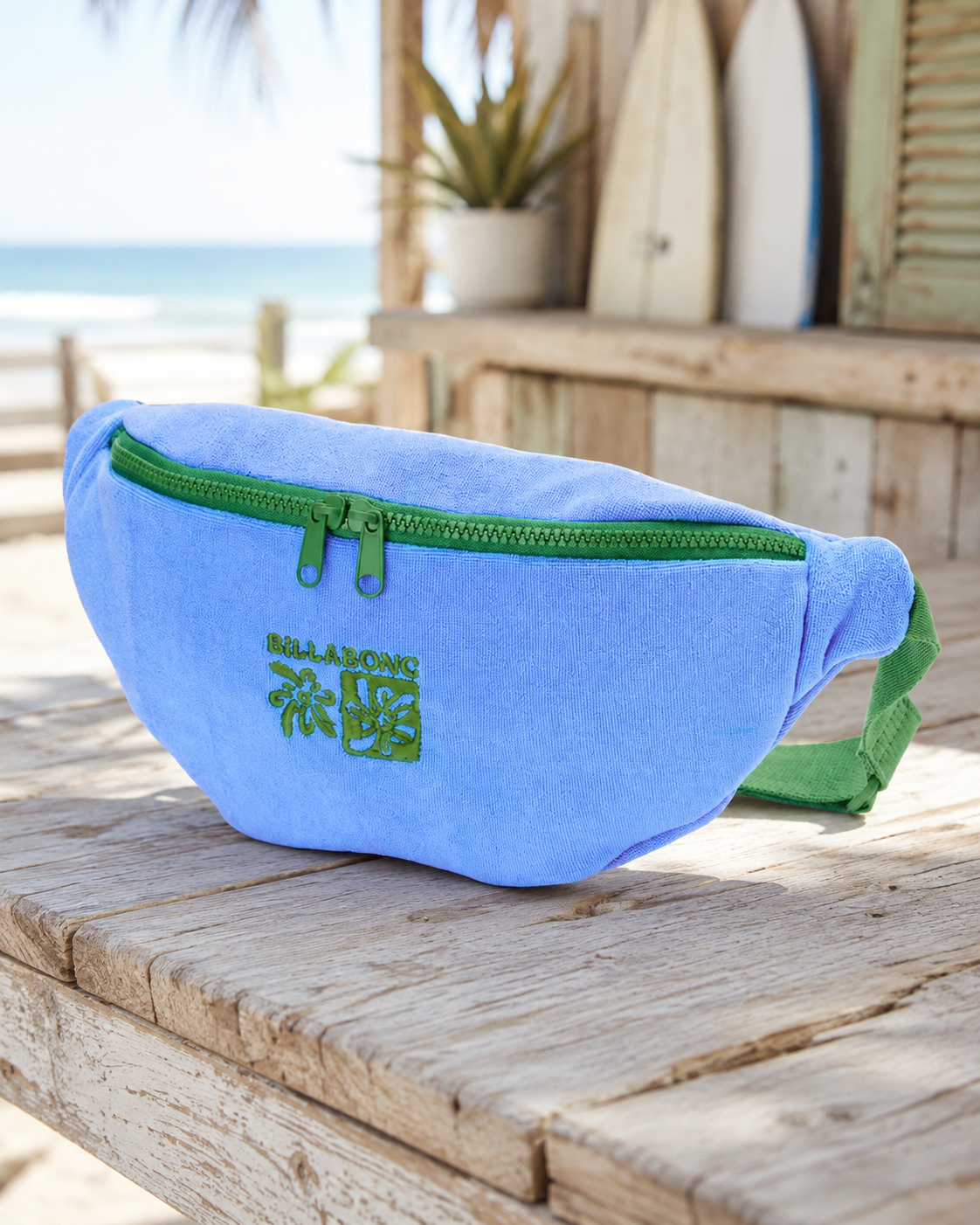 Riñonera Billabong Long Weekend True Blue azul con cremallera verde y logo bordado, bolso de cintura estilo surf lifestyle sobre mesa de madera con ambiente playa.