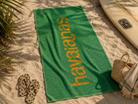 Toalla Havaianas verde y amarilla sobre arena junto a tabla de surf y accesorios beach lifestyle en ambiente premium Costa del Sol